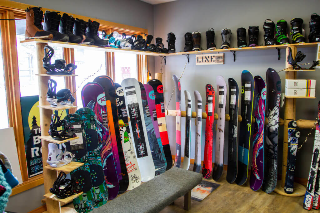 Interior of Ski shop with snowboards lined up on display and a higher shelf with ski and snowboard boots and bindings.