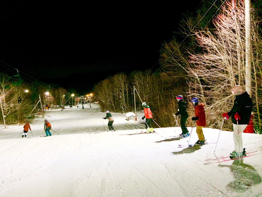 A line of women skiiers start to descend down a slope lit up with led lights.