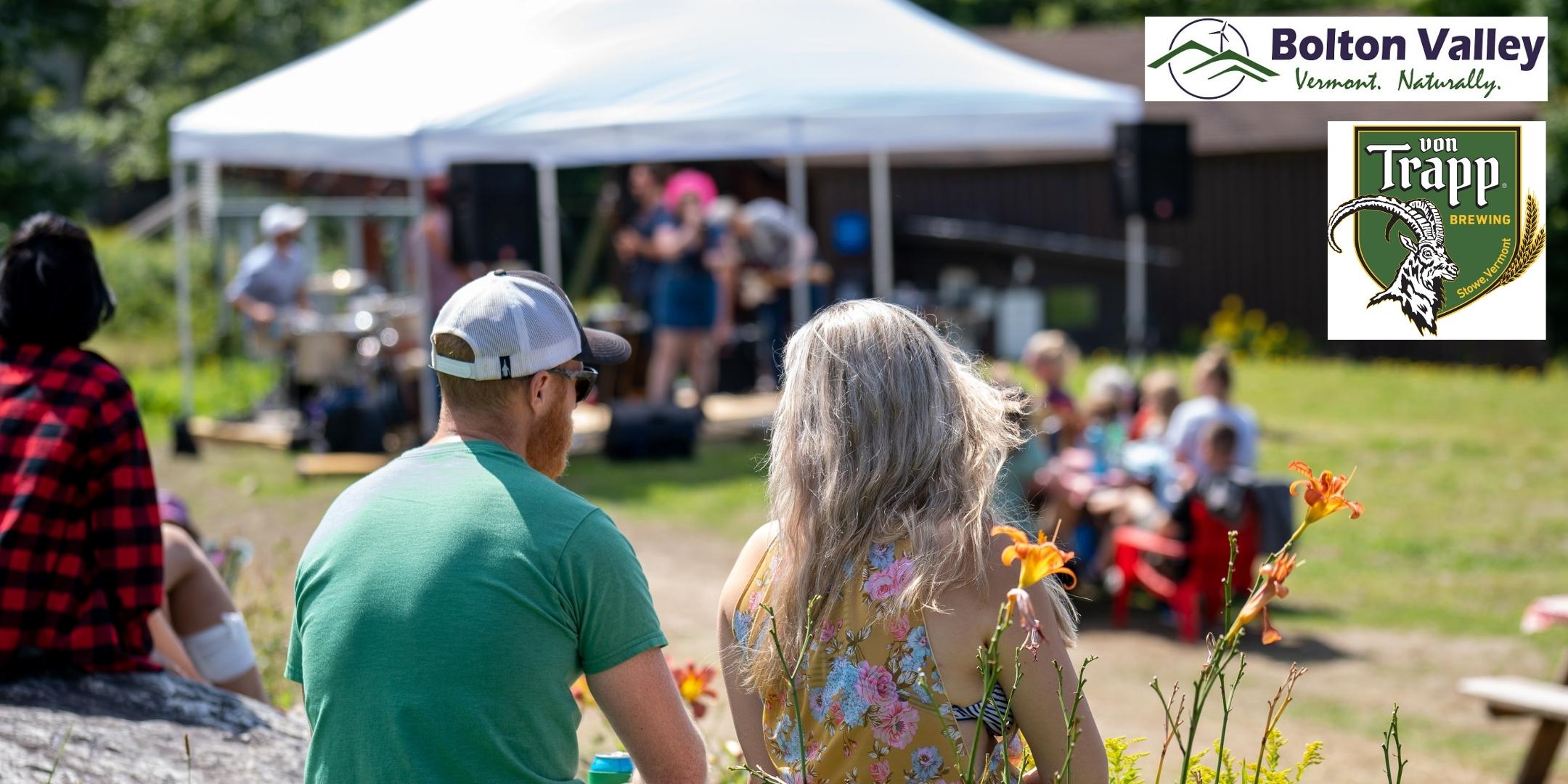 Two people sit on lawn watching live music under a tent on a sunny day.