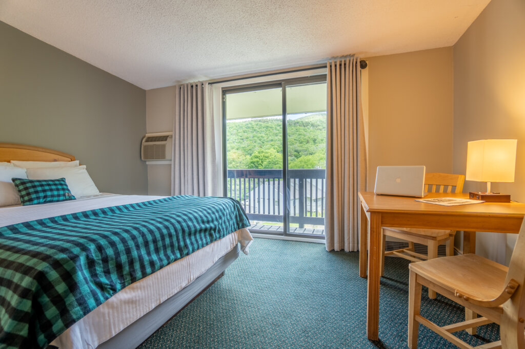 Interior of hotel room with bed, table, chairs, lamp, and balcony overlooking green mountains