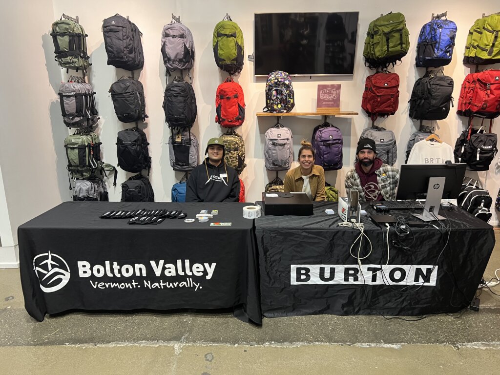 Three Bolton Valley employees sit behind a wall of back packs in front of a table with cloths branded Bolton Valley and Burton