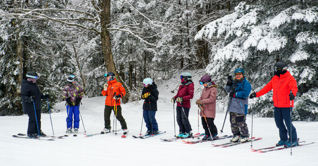 group of skiers line up on snow in front of snow covered trees